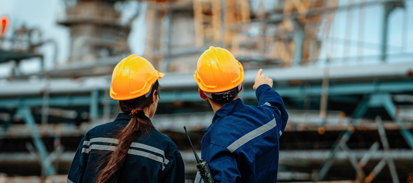 Two workers wearing hard hats examine machinery at an industrial facility. One points toward equipment while discussing safety protocols, emphasizing teamwork and attention to detail.